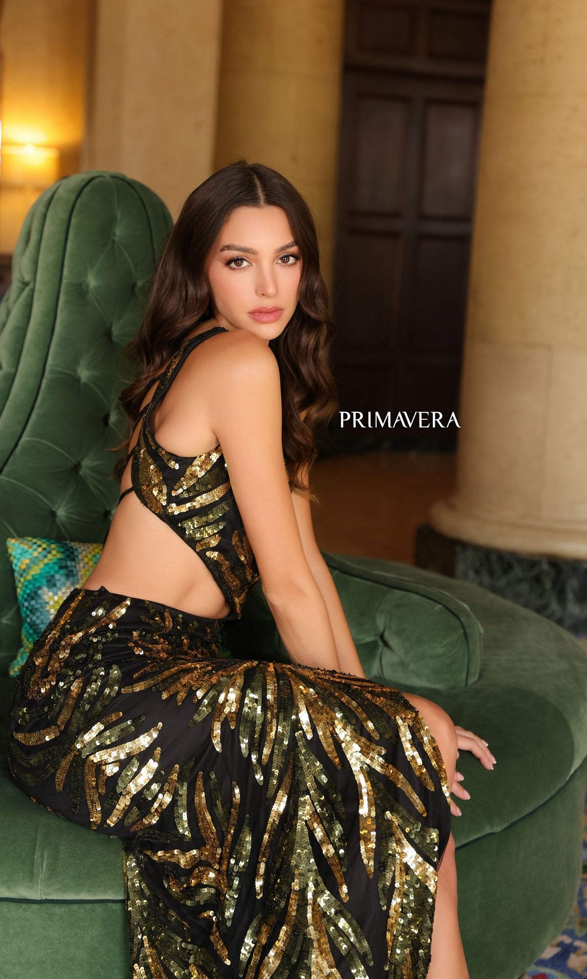 A woman in the One-Shoulder Primavera Long Beaded Prom Dress 4119, featuring black and gold beads, sits on a green armchair in an elegant room with columns;